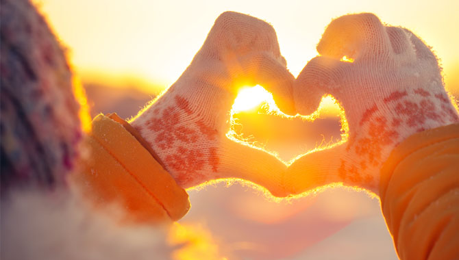 Winterlich gekleidete Person umrahmt Sonne mit zu Herz geformten Fingern